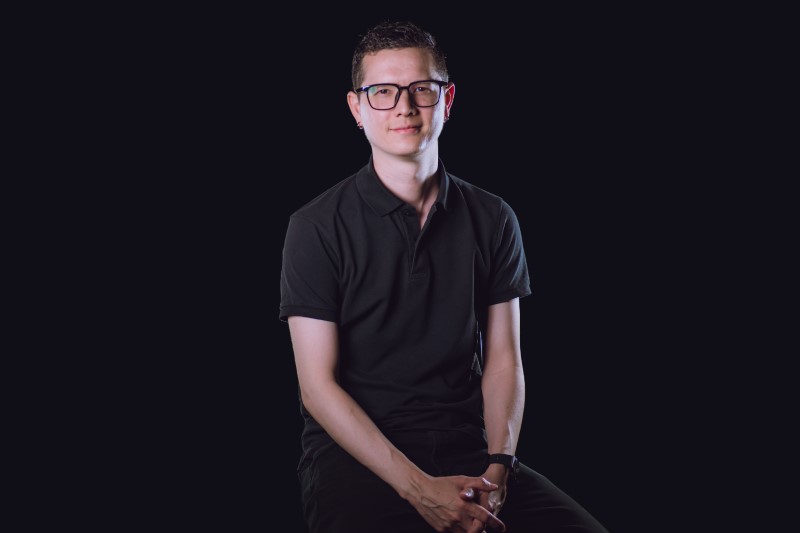 man with dark hair and black-rimmed glasses sitting with hands joined in front of him, wearing black trousers and black polo top, against a black background